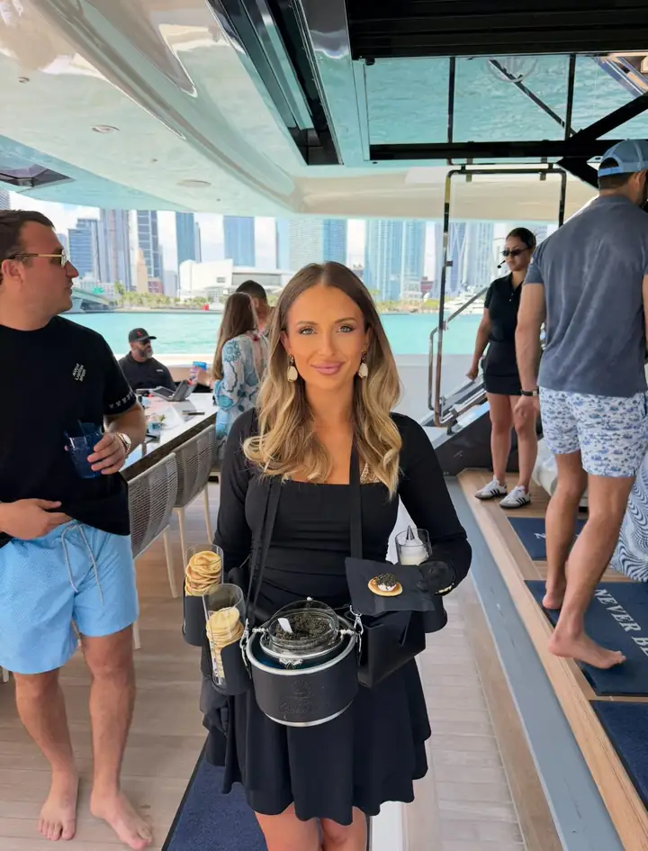An attendant on a yacht deck holds a finished blini, the city skyline behind