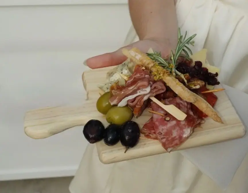 A guest holds a small board of prosciutto, salami, olives, cheeses and a breadstick with rosemary