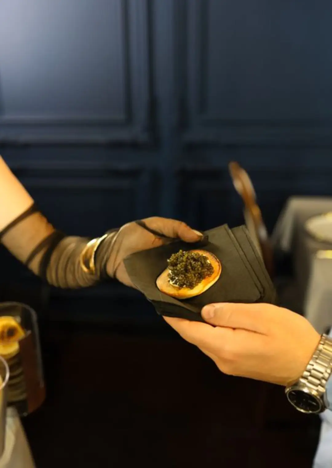 A sheer-gloved attendant hands a guest a blini topped with caviar, on a black napkin, against a navy panelled wall